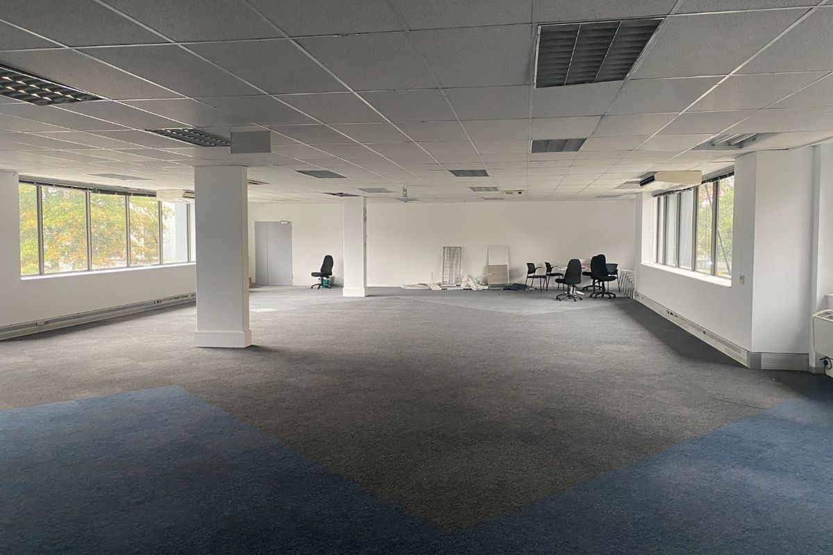 Spacious open-plan commercial office area featuring large windows, suspended ceilings with recessed lighting, and carpeted flooring.