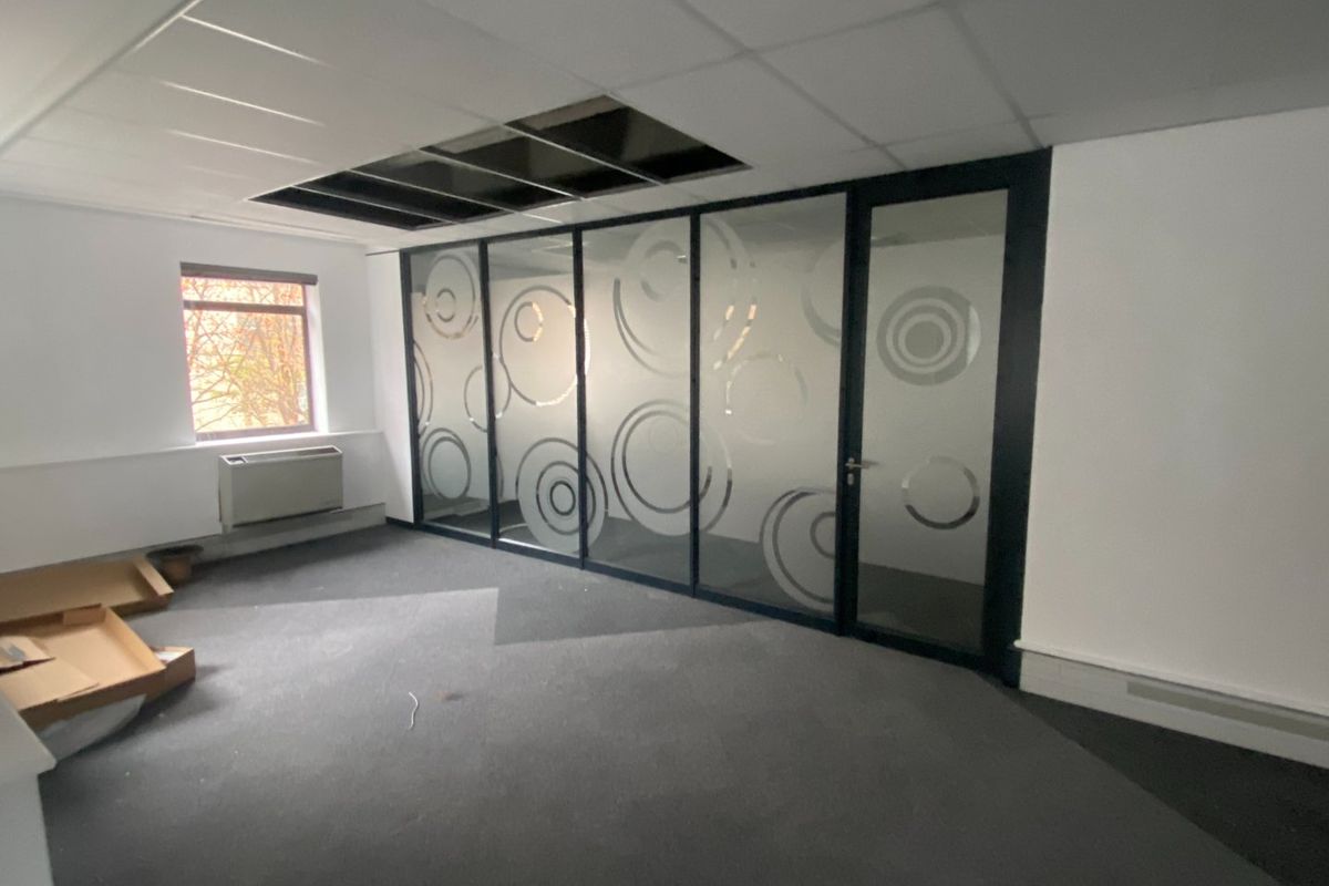 Empty office space featuring a glass partition wall, carpeted flooring, and a suspended ceiling with missing tiles requiring repair.