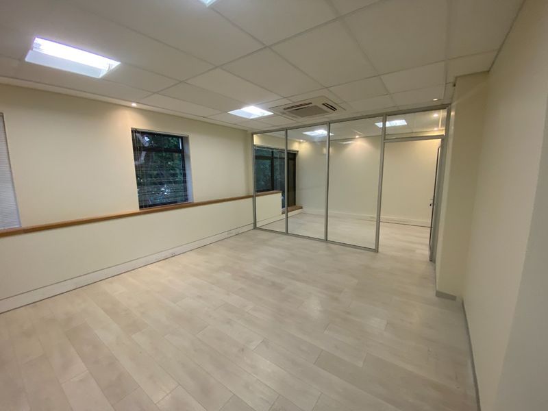A bright, vacant commercial office space featuring modern laminate flooring, a glass partition wall, recessed ceiling lighting, and integrated air conditioning.