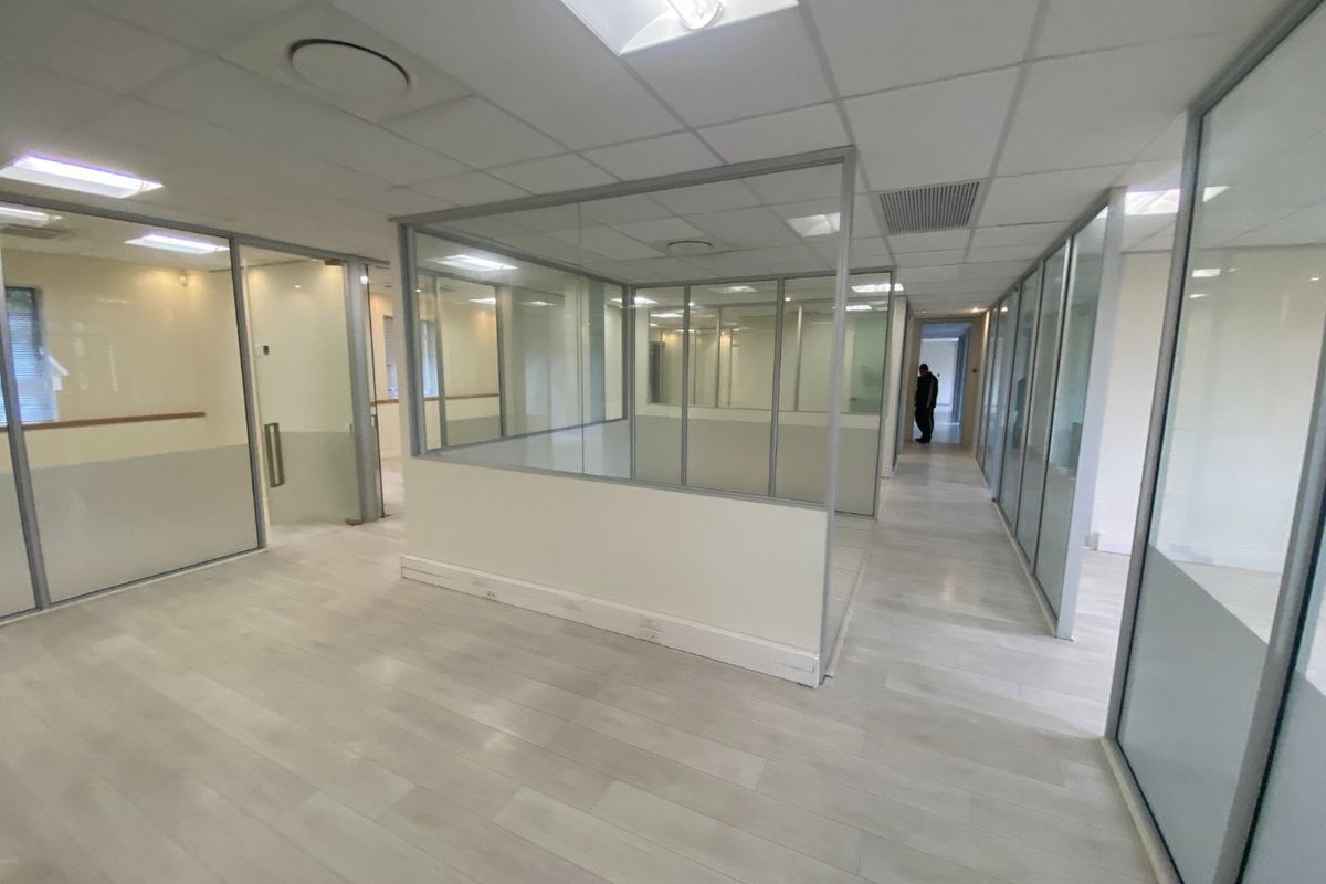 Modern commercial office space featuring glass partition walls, suspended ceilings with recessed lighting, and light-toned laminate flooring in an open-plan layout.