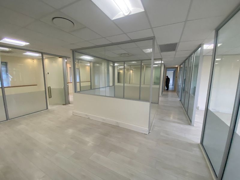Modern commercial office space featuring glass partition walls, suspended ceilings with recessed lighting, and light-toned laminate flooring in an open-plan layout.