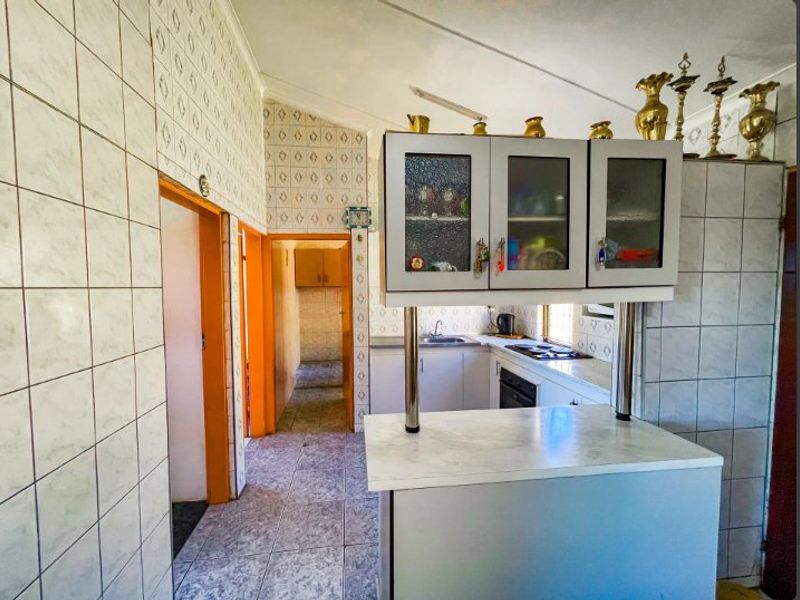 A dated kitchen featuring tiled walls, built-in cabinetry, and a breakfast bar, with an adjacent scullery or pantry area visible through the doorway.