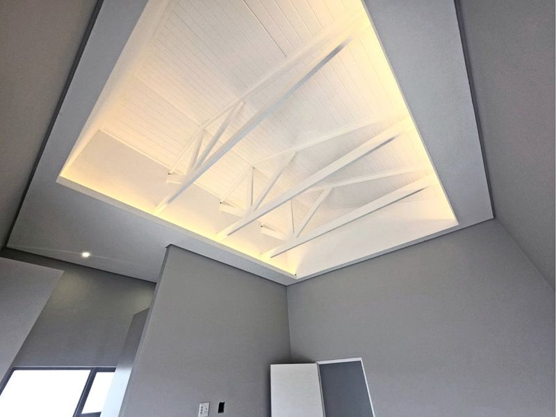 Modern living area featuring a high vaulted ceiling with exposed white timber trusses, recessed cove lighting, and clean grey wall finishes.