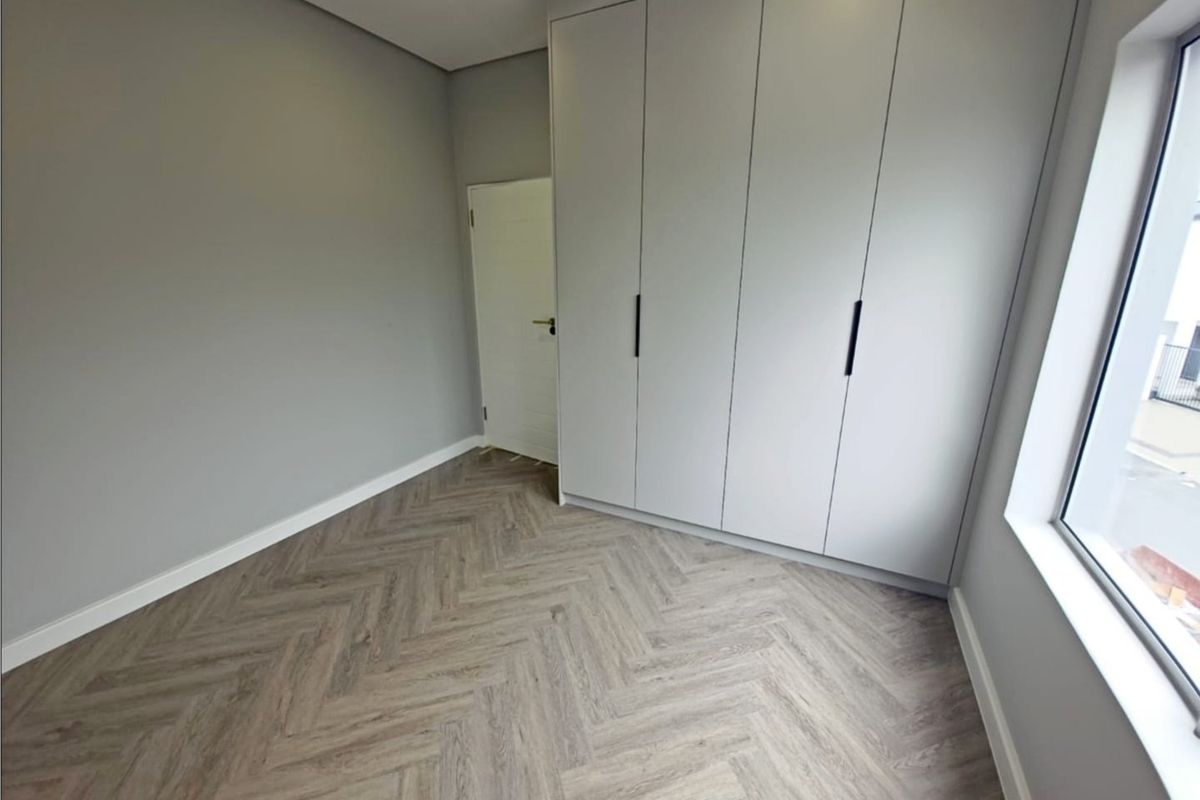 A modern, empty bedroom featuring light grey walls, built-in cupboards, and contemporary herringbone flooring with ample natural light.