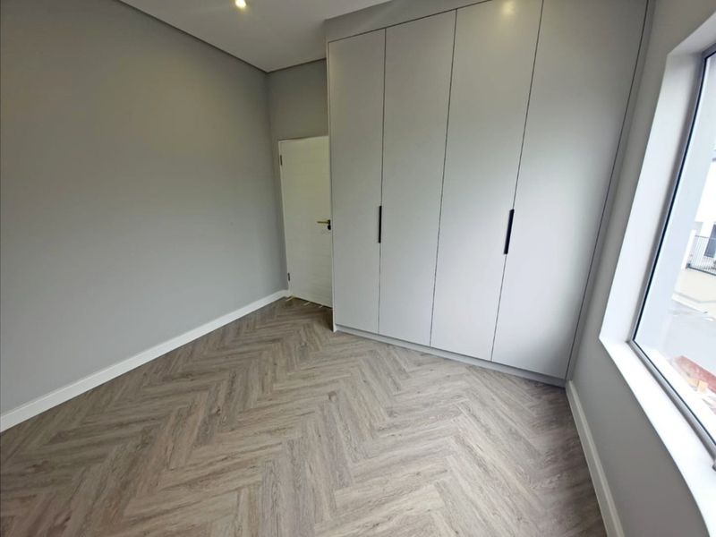 A modern, empty bedroom featuring light grey walls, built-in cupboards, and contemporary herringbone flooring with ample natural light.
