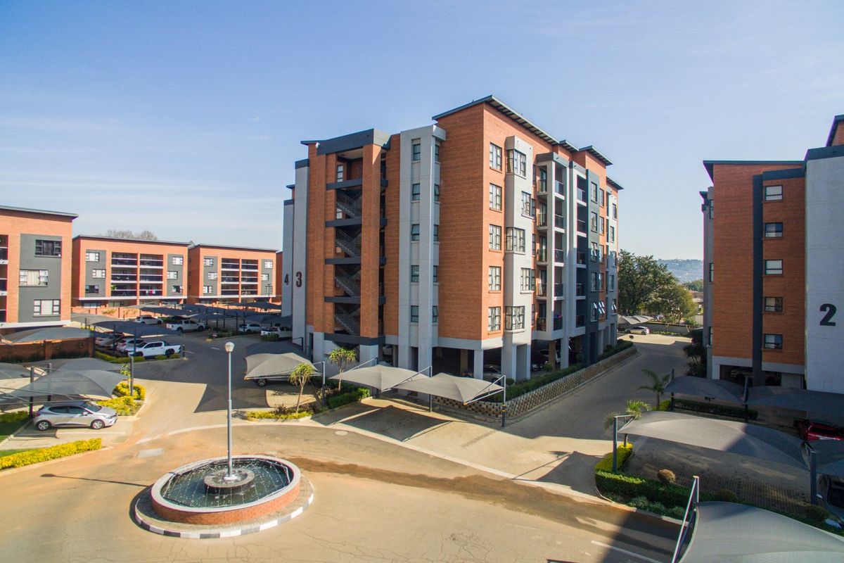 Aerial view of a modern multi-story apartment complex featuring brick facades, balconies, covered parking bays, and a central fountain in the driveway.