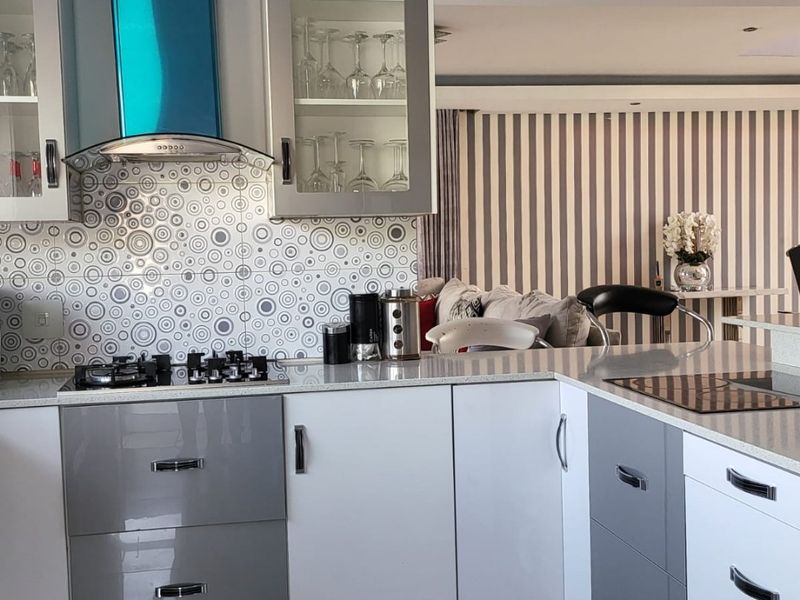 Modern open-plan kitchen featuring high-gloss cabinetry, stone countertops, a gas hob with extractor fan, and decorative tiled backsplash, flowing into the living area.