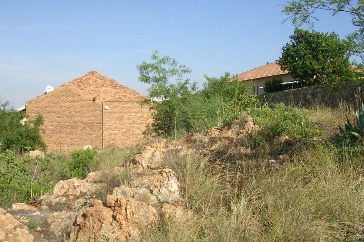 A view of a sloped residential plot featuring natural rock formations, native vegetation, and a brick building structure in the background.