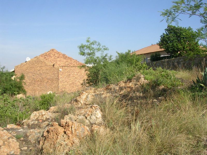 A view of a sloped residential plot featuring natural rock formations, native vegetation, and a brick building structure in the background.