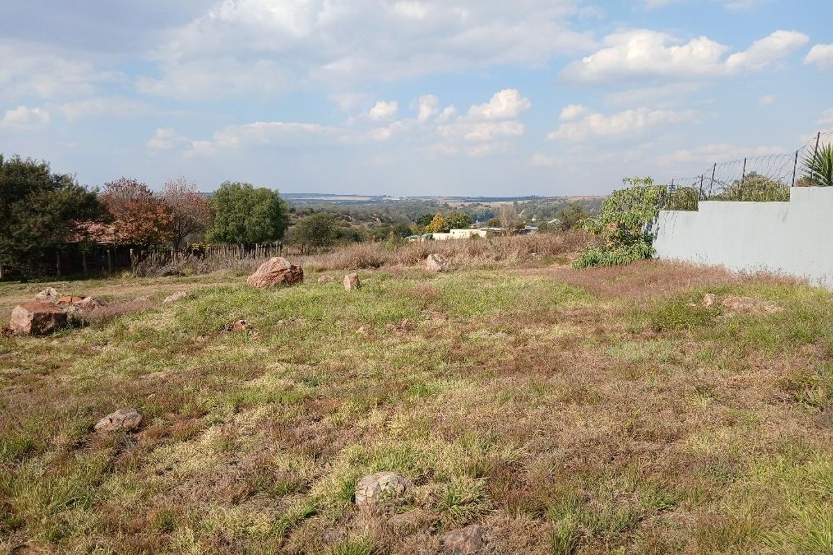 A view of a vacant residential plot featuring natural grass, scattered rocks, and a perimeter wall with security fencing.