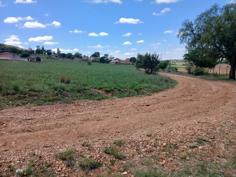 A view of an unpaved access road leading to a vacant residential plot with surrounding grassland and established neighborhood structures in the distance.