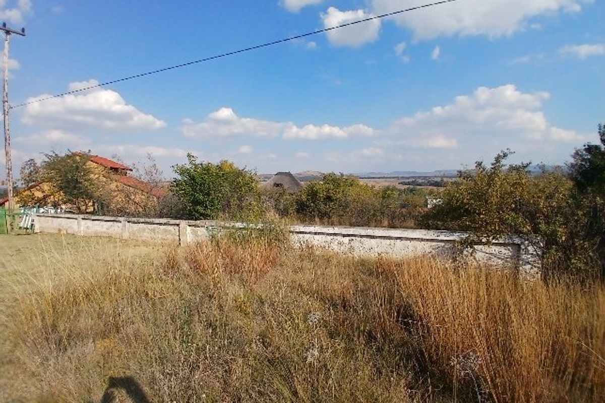 A view of a vacant residential plot featuring a boundary wall, utility pole, and natural vegetation with distant landscape views.