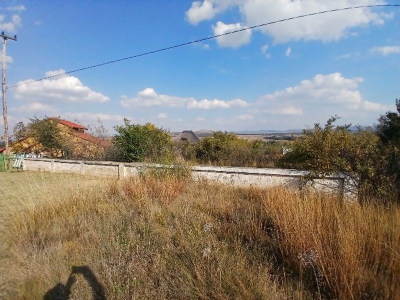 A view of a vacant residential plot featuring a boundary wall, utility pole, and natural vegetation with distant landscape views.