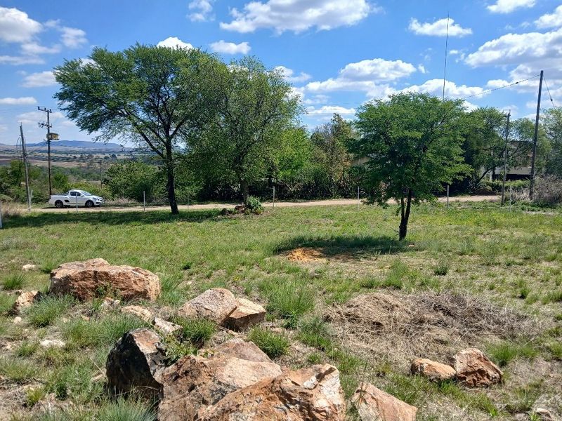 A view of a vacant residential plot featuring natural rock formations, established trees, and perimeter fencing with utility access.