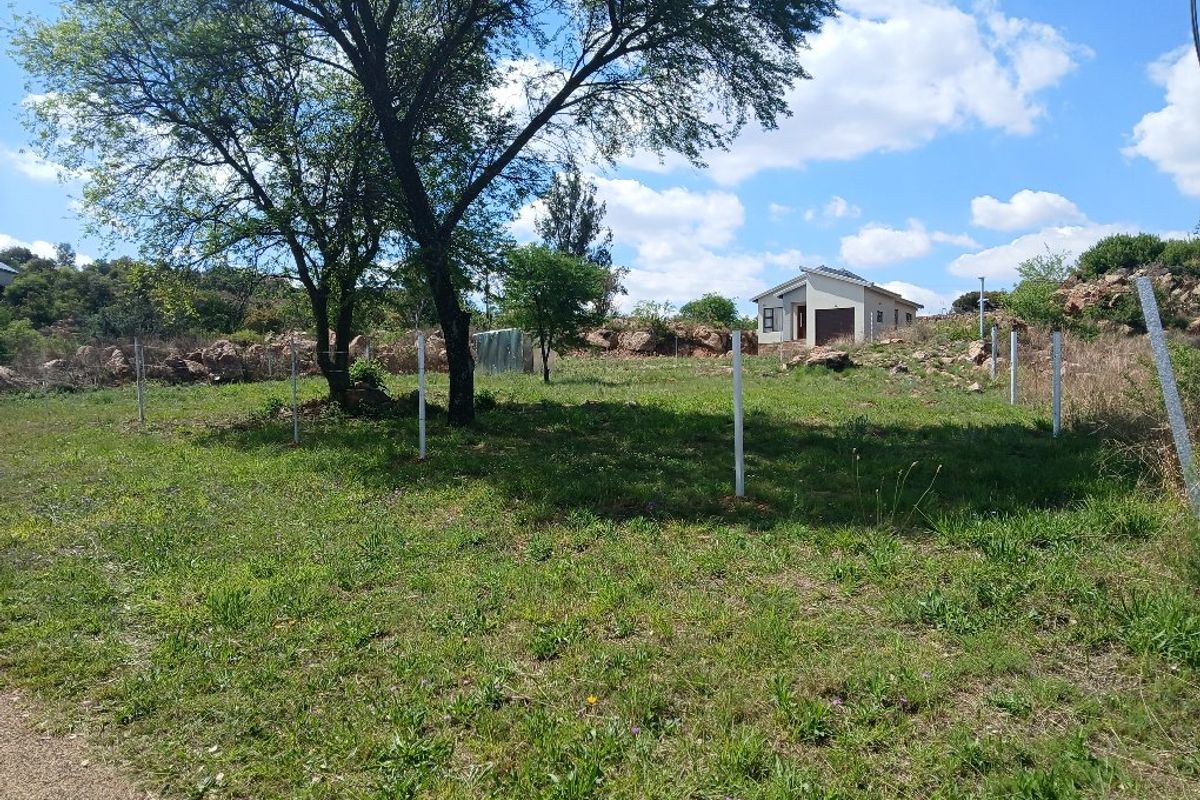 A view of a vacant residential plot featuring established trees, rocky terrain, and perimeter fencing posts, with a neighboring house visible in the background.