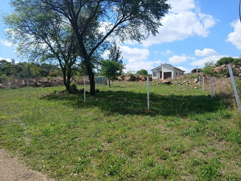 A view of a vacant residential plot featuring established trees, rocky terrain, and perimeter fencing posts, with a neighboring house visible in the background.