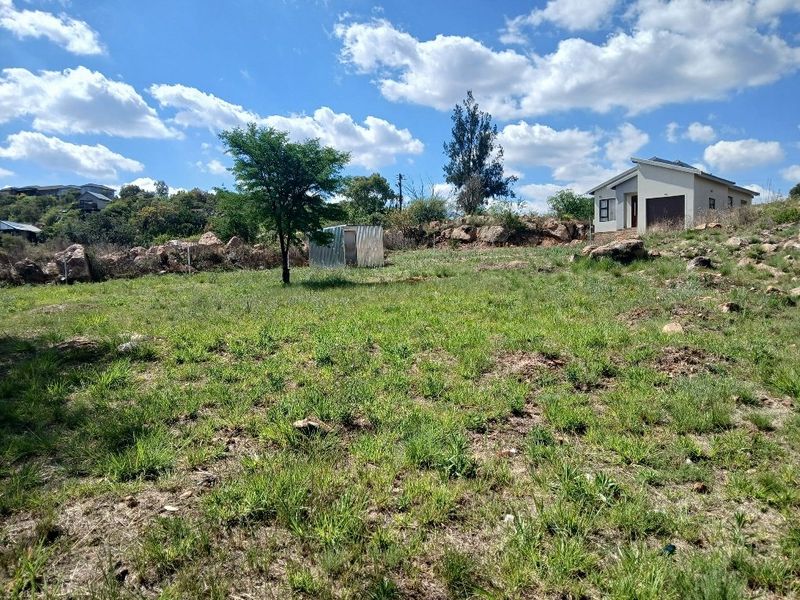 A view of a sloped residential plot featuring natural rock formations, native grass, and a small metal storage shed on the property.