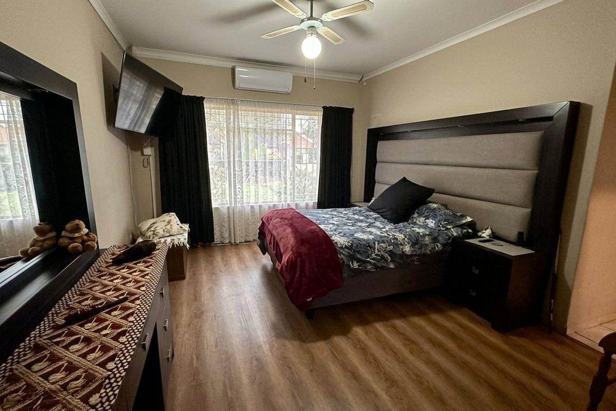 A spacious bedroom featuring laminate flooring, a ceiling fan, and a wall-mounted air conditioning unit, with security bars installed on the window.