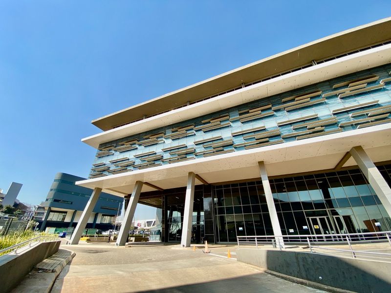 Modern commercial building facade featuring extensive glass curtain walling, integrated sun louvers, and prominent concrete support columns at the main entrance.