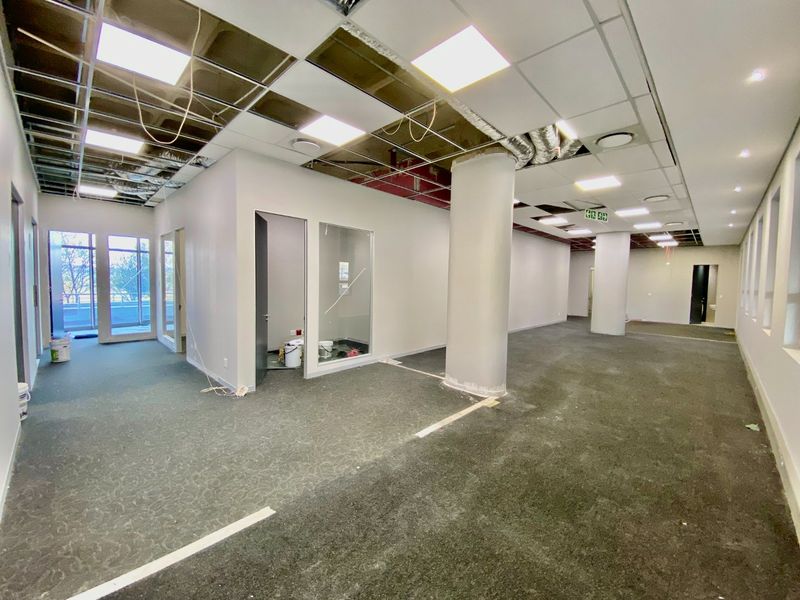 Open-plan commercial office space featuring structural columns, glass partitions, and carpet flooring, currently undergoing ceiling maintenance and repairs.