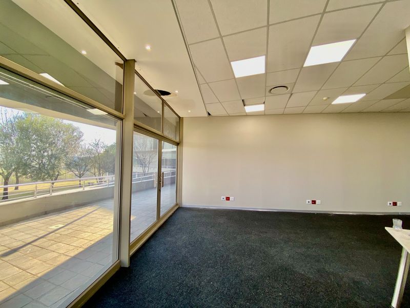 Empty commercial office space featuring large glass sliding doors leading to a balcony, carpeted flooring, and a suspended ceiling with recessed lighting.
