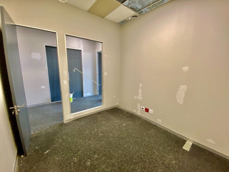 An empty commercial office space featuring a glass partition, carpeted flooring, and a suspended ceiling, currently requiring wall repairs and ceiling maintenance.