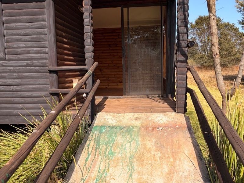 Exterior view of a log cabin featuring a wooden deck, sliding glass door, and a concrete access ramp with visible weathering.