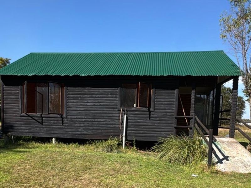 A detached timber log cabin featuring a green corrugated metal roof and a wooden entrance ramp, set within a grassy outdoor area.