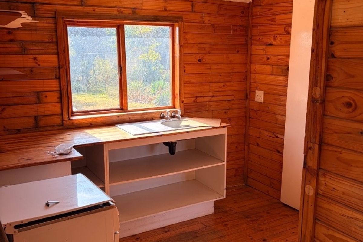 A rustic kitchen area featuring timber wall cladding, wooden flooring, a built-in sink unit with open shelving, and natural light from a window.