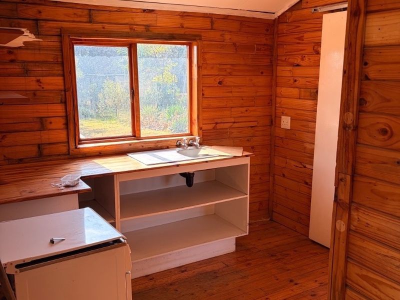 A rustic kitchen area featuring timber wall cladding, wooden flooring, a built-in sink unit with open shelving, and natural light from a window.