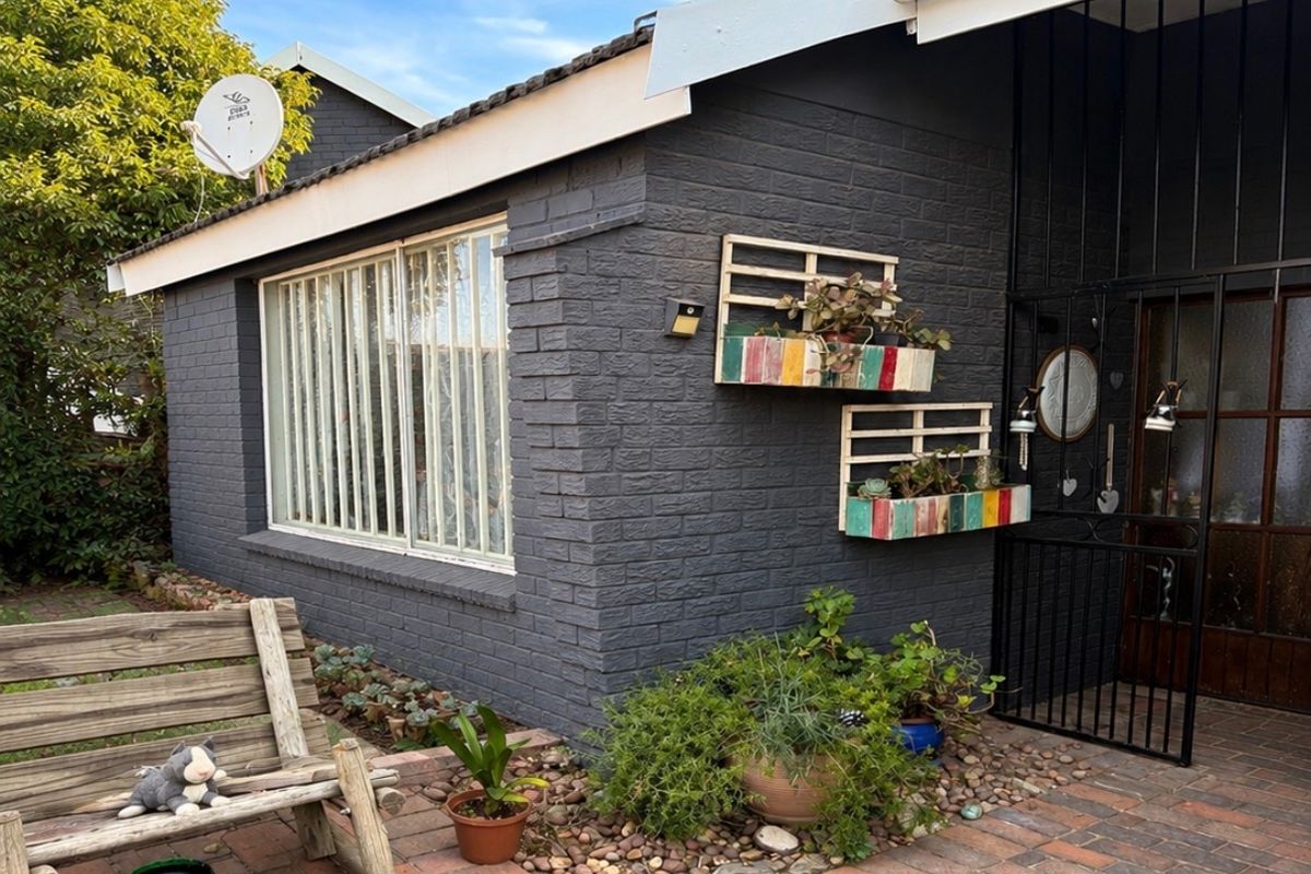 A well-maintained rear exterior view featuring dark painted brickwork, a paved patio area, and secure metal entrance gate.