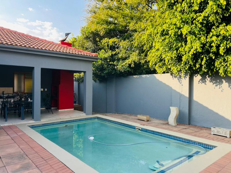 Outdoor pool area featuring a covered patio with a built-in braai, surrounded by a private perimeter wall and mature trees.