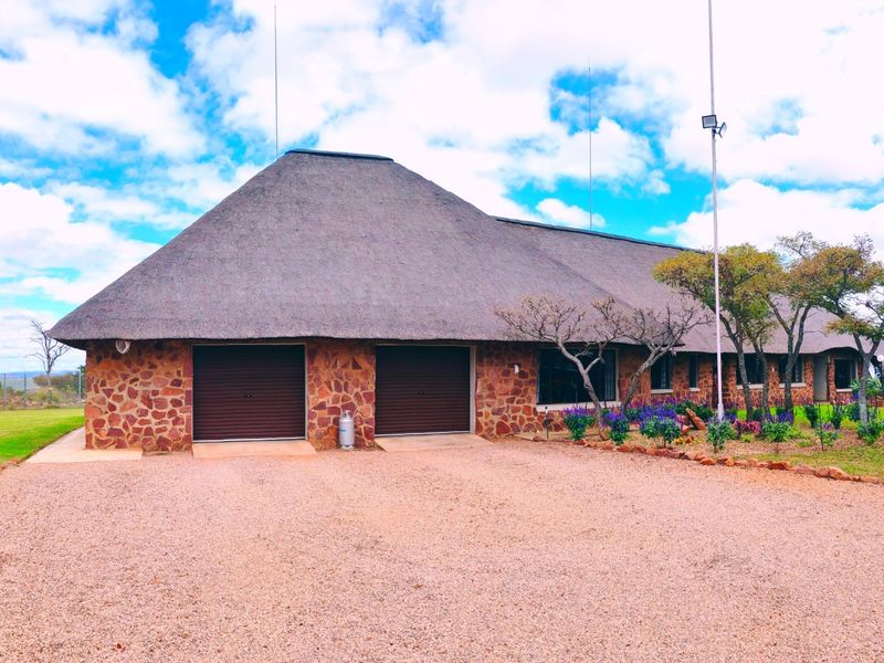 Front view of a farm-style residence featuring a traditional thatch roof, stone exterior cladding, and a double garage, set on a spacious gravel driveway.