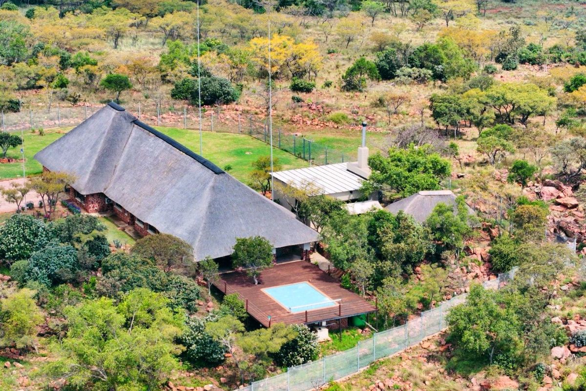 Aerial view of a farm property featuring a large thatch-roof dwelling, an elevated timber deck with a swimming pool, and surrounding natural bushveld landscape.