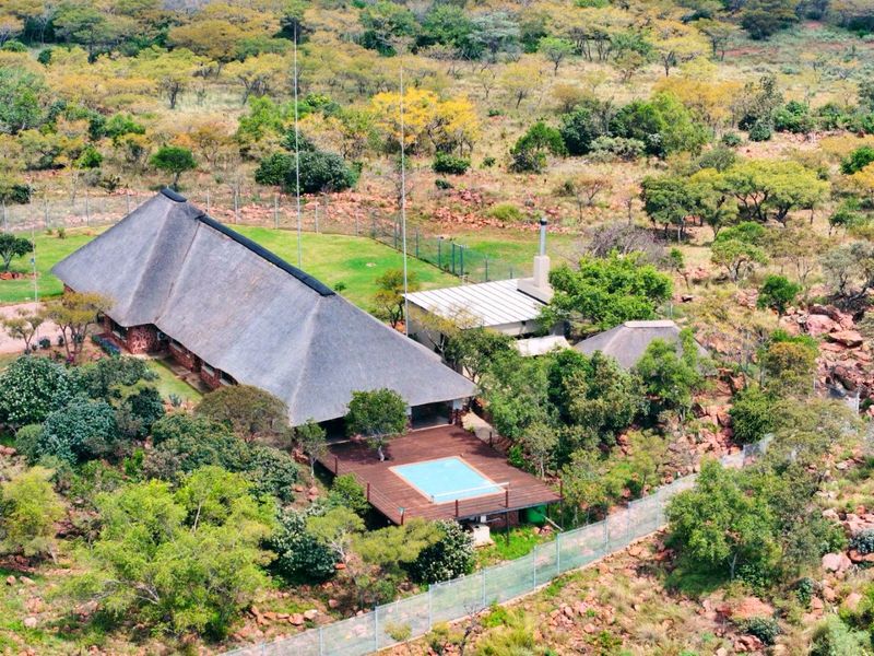 Aerial view of a farm property featuring a large thatch-roof dwelling, an elevated timber deck with a swimming pool, and surrounding natural bushveld landscape.