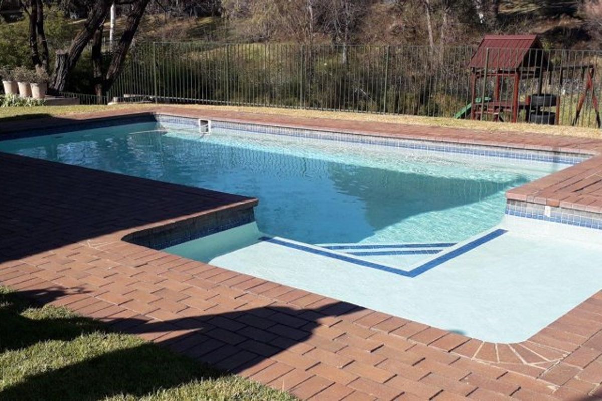 A well-maintained swimming pool area featuring brick paving surround and a secure metal perimeter fence, set against a natural backdrop.