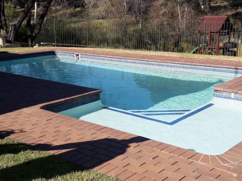 A well-maintained swimming pool area featuring brick paving surround and a secure metal perimeter fence, set against a natural backdrop.