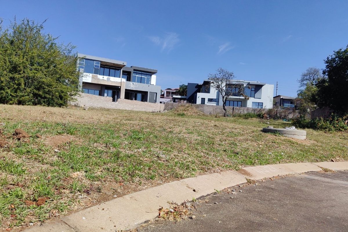 A vacant residential plot of land with a gentle slope, situated in a developed neighborhood with modern homes visible in the background.