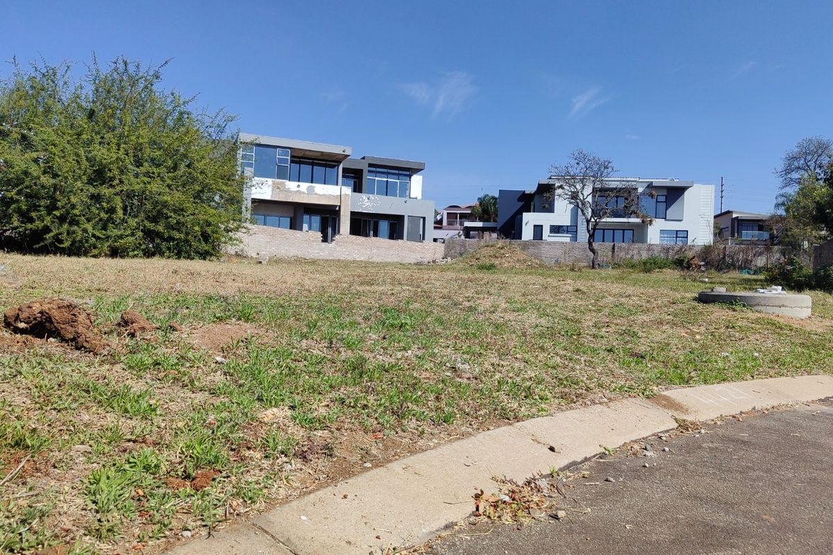 A vacant residential plot featuring a gentle slope, concrete curbing, and established neighboring modern homes in a developed estate setting.