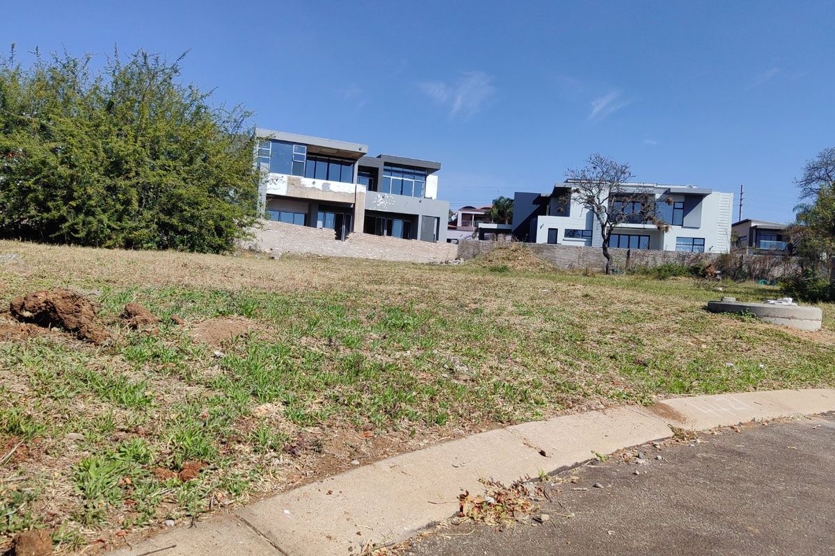 A vacant residential plot of land featuring a gentle slope, bordering an asphalt road with a concrete curb and established neighboring properties.