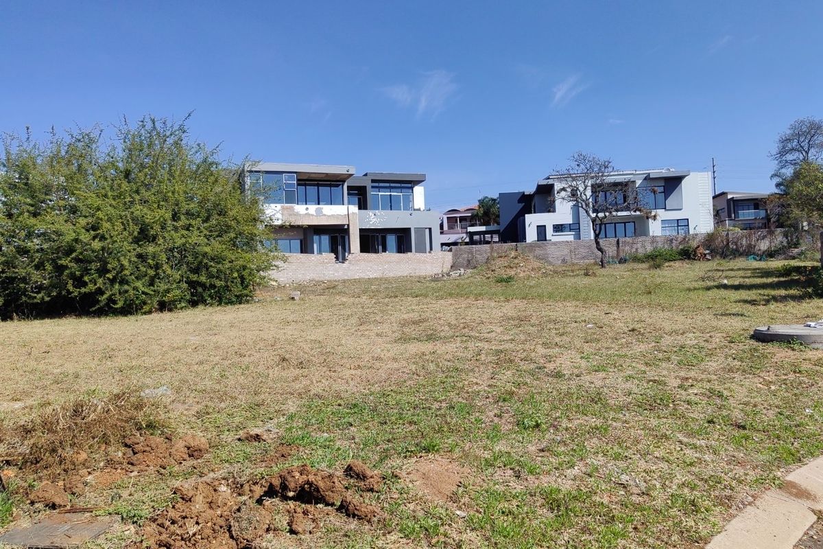 A vacant residential plot of land featuring natural grass terrain, a retaining wall, and views of neighboring modern multi-story homes.