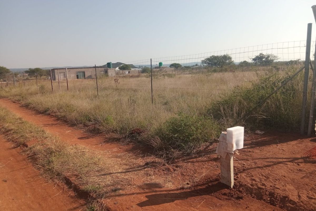 A vacant residential plot featuring wire mesh perimeter fencing and direct access from a dirt road, situated in a developing residential area.