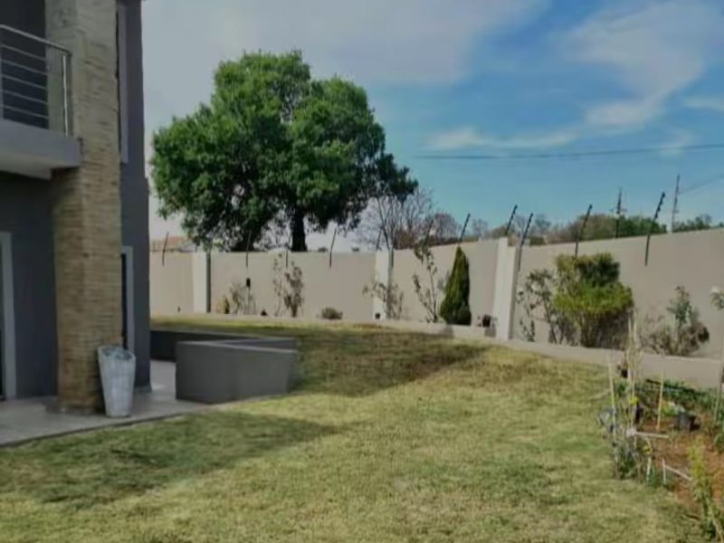 A spacious rear garden featuring a well-maintained lawn, perimeter wall with electric fencing, and a stone-clad pillar supporting an upper-level balcony.