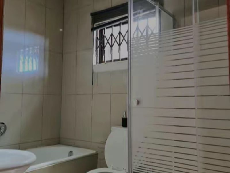 A functional bathroom featuring a bathtub, separate shower enclosure, toilet, and washbasin with neutral wall tiling and security bars on the window.