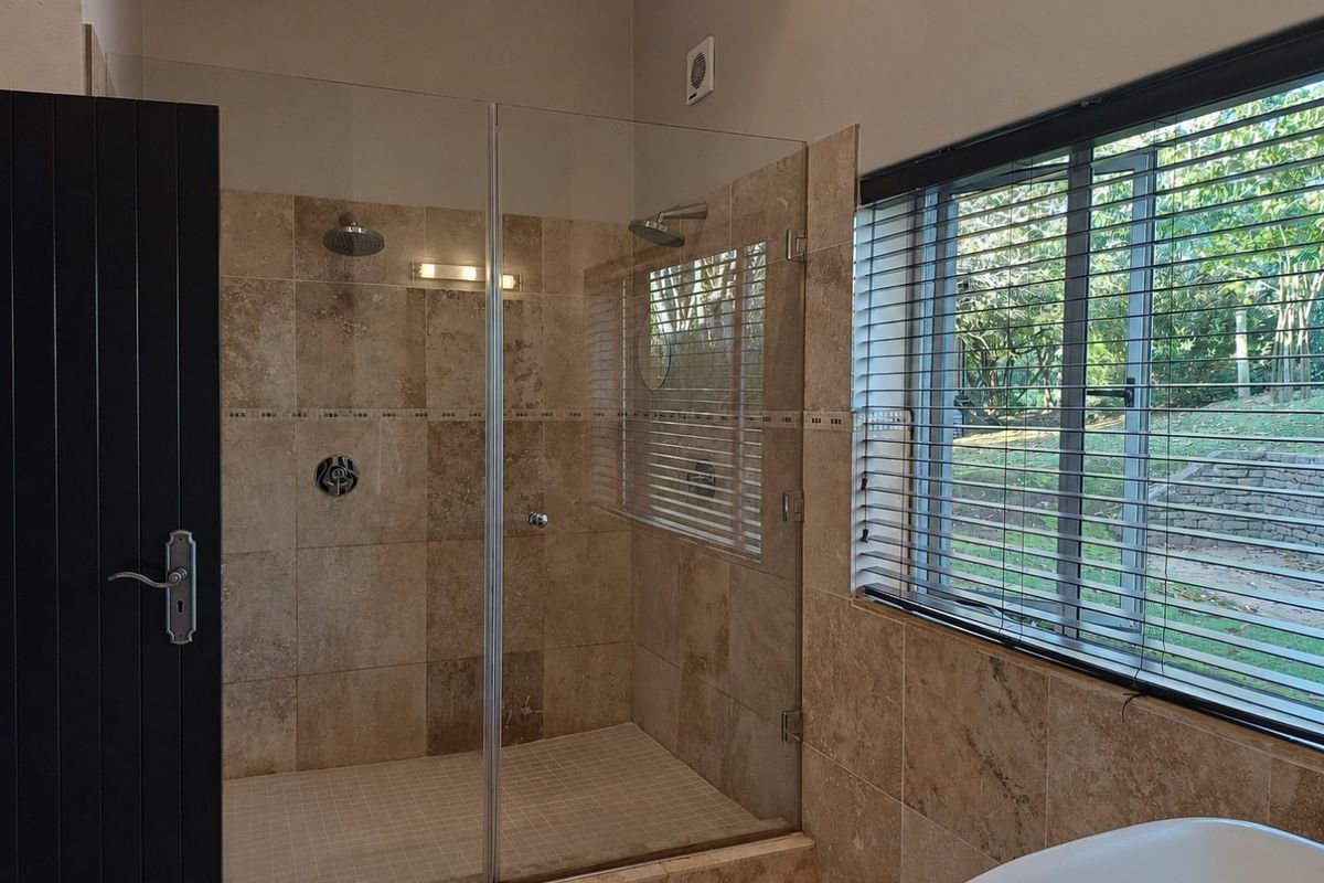 Modern bathroom featuring a spacious walk-in shower with travertine tiling, rainfall shower head, and large window with venetian blinds.