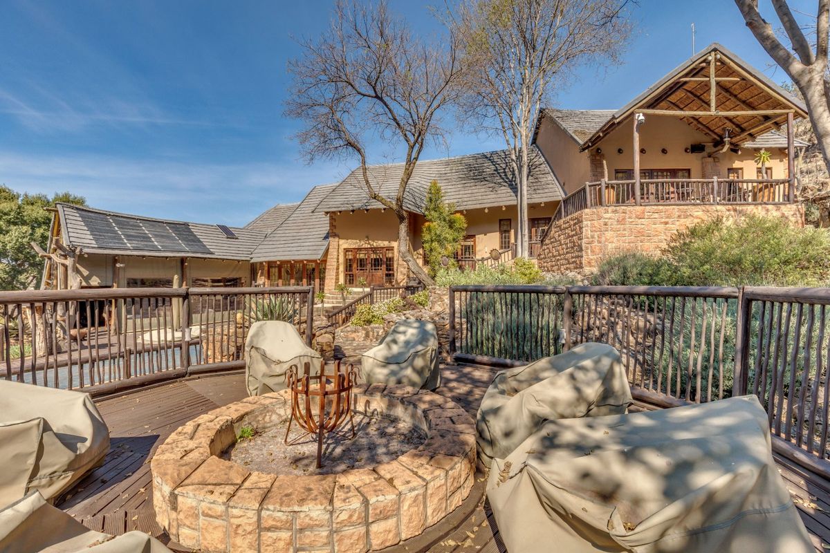 A spacious rear outdoor entertainment area featuring a stone fire pit, timber decking, and views of the main residence with stone and thatch architecture.