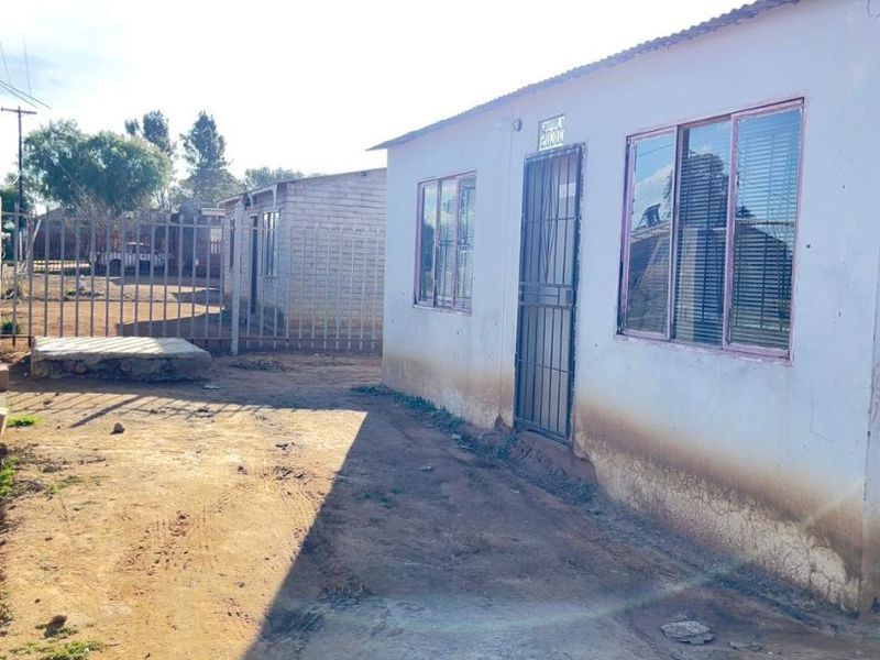 Exterior view of a single-story house featuring metal window frames, a security gate, and a corrugated metal roof, set on an unpaved plot.