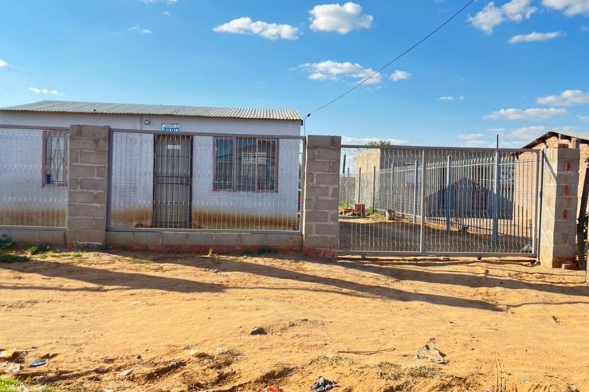 Front exterior view of a single-story house featuring a corrugated metal roof, concrete block perimeter wall, and metal security gate and door.