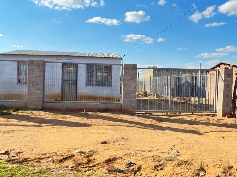 Front exterior view of a single-story house featuring a corrugated metal roof, concrete block perimeter wall, and metal security gate and door.