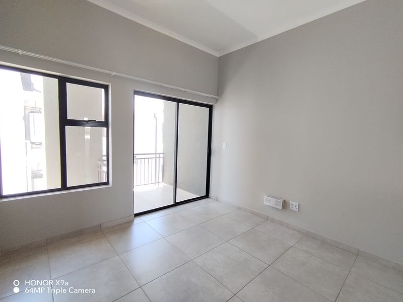 A bright, empty living area featuring neutral tiled flooring, large aluminium-framed windows, and a sliding door leading to an outdoor balcony.